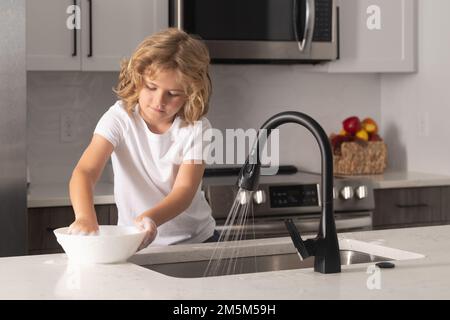 Il bambino con la spugna con il liquido di lavaggio del piatto sta facendo i piatti nella cucina domestica usando la spugna di lavaggio e la lavastoviglie. Pulizia lavastoviglie lavello cucina Foto Stock