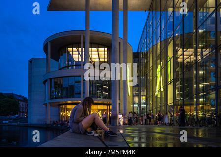 Giovane donna seduta sul lato di Spree accanto all'edificio Paul Loebe nel quartiere governativo di Berlino durante il tramonto Foto Stock
