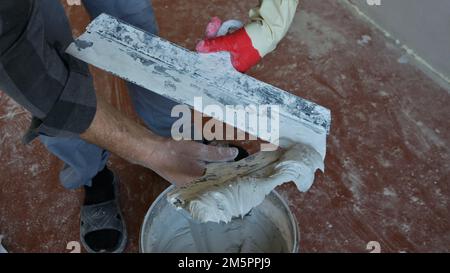 le mani del maestro in un guanto tengono due cazzoletti con un mortaio spesso grigio sopra un contenitore, preparando un materiale di lavoro per le superfici intonacate Foto Stock