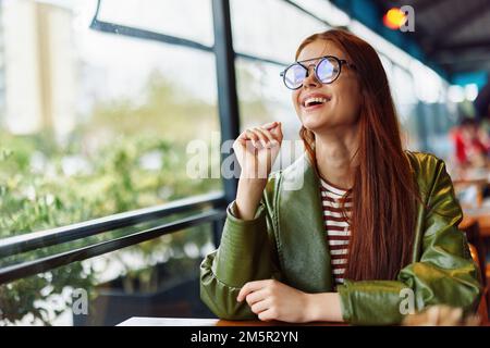 Bella donna con i capelli rossi e bicchieri siede in città in un caffè e guarda fuori la finestra sorridere con i denti, donna freelance blogger Foto Stock