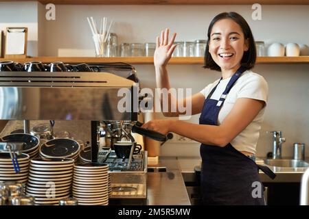 Barista asiatico entusiasta, ragazza che usa la macchina del caffè, che sventola al cliente, salutando gli ospiti nel suo caffè Foto Stock