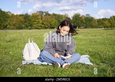 Ritratto di ragazza asiatica lettura libro, seduta sulla sua coperta nel parco, con erba verde, sorridendo felicemente Foto Stock