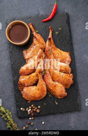 Prelibatezze. Cosce di quaglia grigliate alla griglia in marinata e spezie su ardesia nera con erbe fresche e salsa su sfondo scuro. Alimentazione sana. Vista dall'alto. Foto Stock