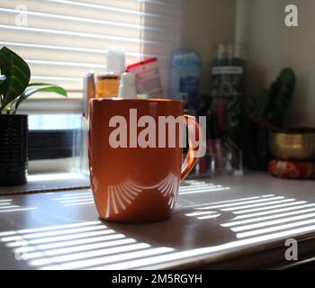 Shot medio di una tazza di caffè arancione con la luce del sole che splende attraverso le persiane Foto Stock