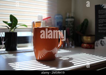 Shot medio di una tazza di caffè arancione con la luce del sole che splende attraverso le persiane Foto Stock