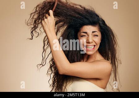 PROBLEMI DI PERDITA DEI CAPELLI. CONCETTO DI CAPELLI LUNGHI. Tormentata urlando infelice impressionante riccio signora latina con spazzola di capelli soffrono con i capelli naughty che posano isolato sopra Foto Stock