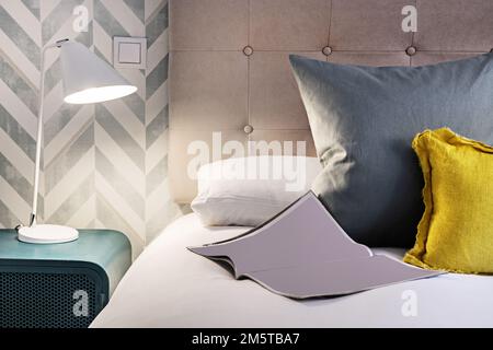 Particolare decorativo di una testata imbottita di un letto in una camera da letto con diversi cuscini, una lampada da comodino bianca e un tavolo in metallo blu Foto Stock