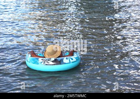 Una donna abbronzante copriva il mio cappello di paglia rilassandosi sul galleggiante gonfiabile del tubo nel mare, nel lago, nel fiume o nella piscina Foto Stock
