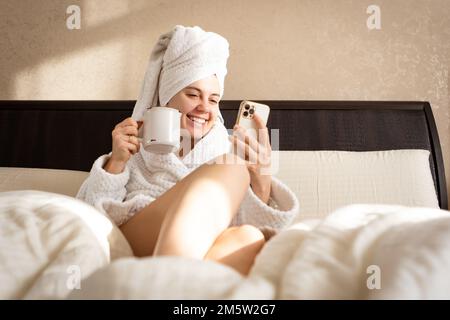 Seduta su un letto in un vestito bianco con un asciugamano sopra la testa è una donna con tazza e mobile Foto Stock