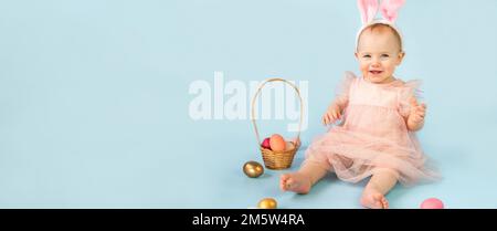 Ragazza felice, dieci mesi, con archetto per orecchie conigliate, seduta su sfondo azzurro dello studio e giocando con uova di pasqua colorate. Piccolo Chi Foto Stock