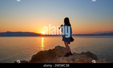 Alba, riflesso del sole sul mare, cielo azzurro-arancio senza nuvole, ragazza da dietro con giacca jeans e macchina fotografica in mano, penisola Rodopou, Gramvoussa Foto Stock