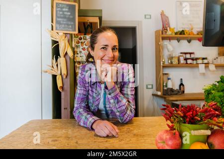 Bella ragazza greengrocer che lavora in negozio appoggiandosi sul banco e circondato da frutta e verdura biologica - Ritratto di giovane imprenditrice donna Foto Stock