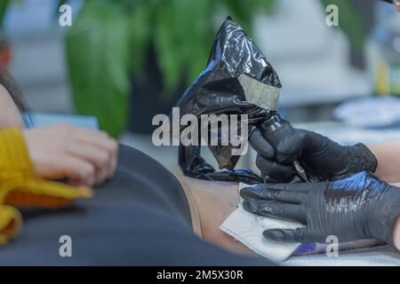 Artista tatuaggio al lavoro. Mani visibili in guanti neri che tengono la pistola da tatuaggio con ago e tracciano una linea sulla parte superiore del braccio del cliente. Foto Stock