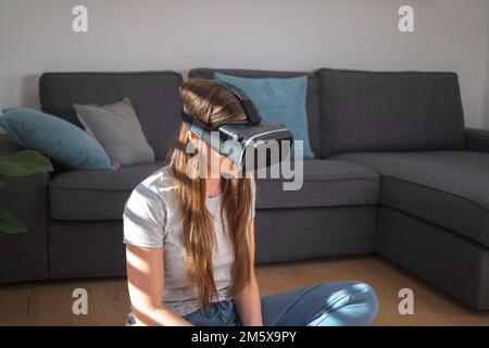 Ragazza seduta sul pavimento del soggiorno, indossando un visore VR e muovendo la mano sullo schermo VR. Mondo virtuale e concetto di realtà aumentata. Foto Stock