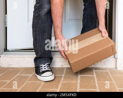 Uomo che raccoglie una scatola di imballaggio consegnata a una porta residenziale. Ordine online consegna pacchetto al portico anteriore di casa. Foto Stock