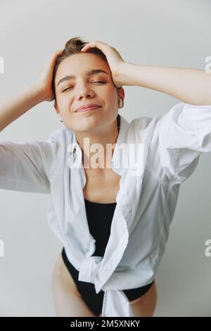 Giovane adolescente donna che ascolta musica con cuffie infertili e ballare a casa, grignando con i denti con un taglio di capelli corto in una camicia bianca su un Foto Stock