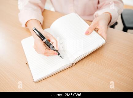 Primo piano delle mani di una donna che scrive in un blocco note posto su un tavolo di legno, ufficio domestico e concetto di lavoro, piani e previsioni Foto Stock