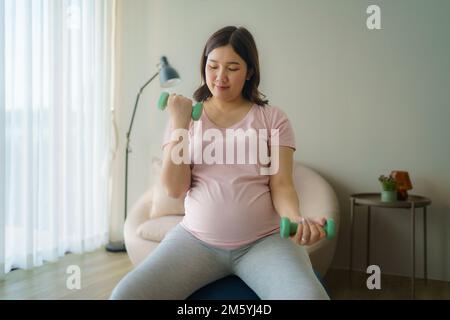 Bella donna asiatica incinta fare esercizio con manubri in soggiorno a casa, concetto sano Foto Stock