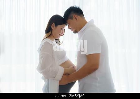 Felice coppia asiatica in attesa di bambino in piedi insieme contro la finestra a casa, amorevole marito teneramente toccando il ventre della sua moglie incinta Foto Stock