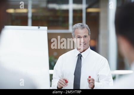 Guida del team. Un uomo d'affari anziano in piedi accanto a una lavagna a fogli mobili con un marcatore in mano e parla con i suoi colleghi in primo piano. Foto Stock