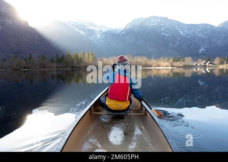 I bambini locali pagaiano in una canoa rossa in una fredda giornata invernale su un tranquillo e bellissimo lago di Bohinj nella regione di Gorenjska in Slovenia Foto Stock