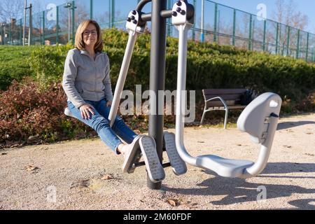 Donna matura che fa esercizi con le gambe in un apparecchio di ginnastica in un parco pubblico. Foto Stock