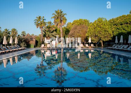 Sedie a sdraio vuote e ombrelloni ripiegati che si riflettono sulla piscina del resort Foto Stock