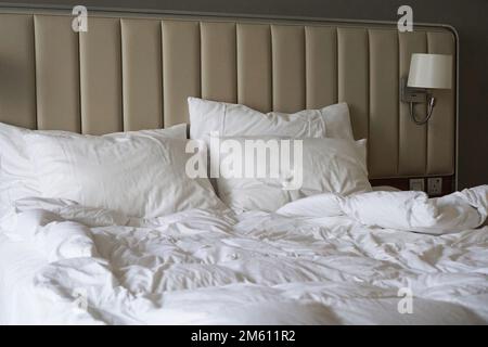 Letto stropicciato in una camera d'albergo o a casa. Lenzuola bianche. Testata imbottita beige. Vista laterale Foto Stock