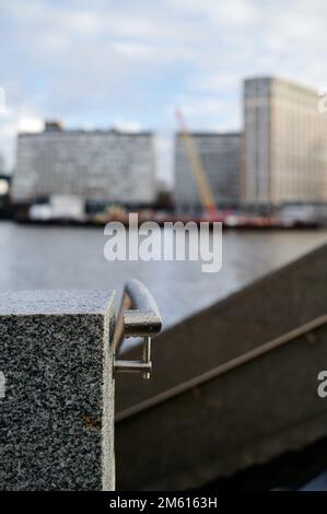 Ringhiera metallica con gocce d'acqua contro il blocco di marmo grigio in un ambiente urbano costruito con sfocatura di fondo degli edifici e del fiume e del cielo Foto Stock