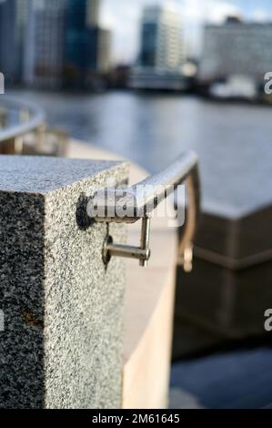 Ringhiera metallica con gocce d'acqua contro il blocco di marmo grigio in un ambiente urbano costruito con sfocatura di sfondo di edifici e fiume Foto Stock