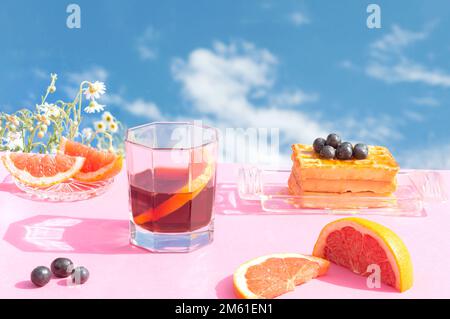 Tavolo per la colazione con succo di frutta, pompelmo, mirtillo, waffle e miele. Fiori e cielo con nuvole sullo sfondo. Estate ancora vita all'aperto co Foto Stock