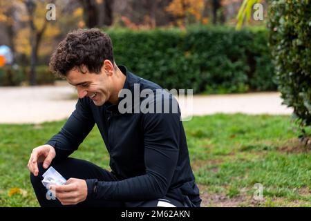 Giovane atleta maschile in abbigliamento sportivo nero seduto sull'erba nel parco in una giornata nuvolosa Foto Stock