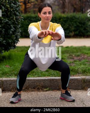 Corpo pieno di atleta giovane e focalizzata in leggings e sneakers squatting sul sentiero vicino a cespugli verdi nel parco Foto Stock