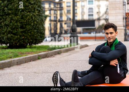 Il ragazzo riccio soddisfatto in abbigliamento sportivo abbraccia le ginocchia mentre si siede in città strada e guardando la macchina fotografica Foto Stock