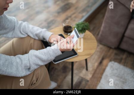 Una donna che acquista online hold carta di credito e tablet digitale mano di giovane donna adulta prendere la decisione di acquisto online mentre si siede a casa re Foto Stock
