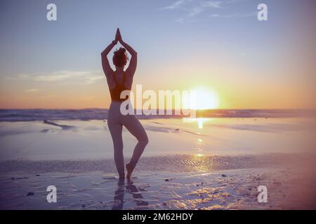 Visto da dietro palestra donna jogger in abiti fitness in spiaggia al tramonto meditando. Foto Stock