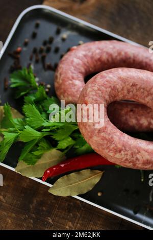 appetitosi prodotti di carne con erbe e verdure. Anelli di salsiccia crudi fatti in casa, prezzemolo fresco, peperoncino rosso in un piatto nero Foto Stock