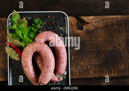 Anelli di salsiccia crudi fatti in casa, prezzemolo fresco, peperoncino rosso in un piatto nero su un tavolo di legno scuro. appetitosi prodotti di carne con erbe e. Foto Stock