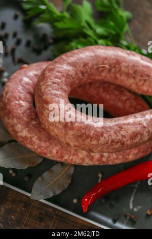 Anelli di salsiccia crudi, prezzemolo fresco, peperoncino rosso in un piatto nero su un tavolo di legno scuro. appetitosi prodotti di carne con erbe e verdure Foto Stock