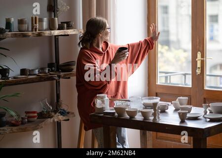 Giovane donna ceramicista bere tè o caffè sul posto di lavoro in un accogliente studio di ceramica Foto Stock