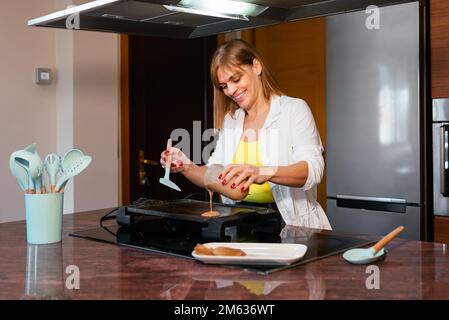 Donna adulta allegra che versa pastella su griglia elettrica calda mentre cucinano frittelle al bancone in cucina moderna Foto Stock