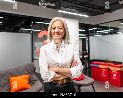 Donna d'affari anziana positiva in vestito di classe in piedi con braccia incrociate in moderno spazio di lavoro durante la pausa dal lavoro in azienda Foto Stock