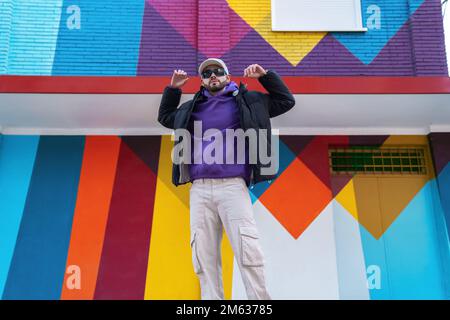 Da sotto maschio in elegante outerwear alzando le braccia e si allunga contro colorato edificio sulla strada cittadina Foto Stock