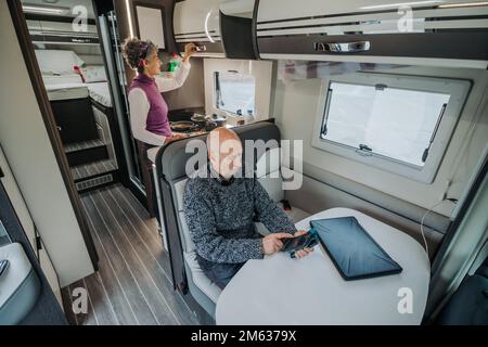 Dall'alto di un uomo anziano in vestito casual seduto al tavolo messaggi sul telefono cellulare, mentre la donna prepara la cena in caravan van Foto Stock