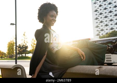 Vista laterale di una splendida giovane ragazza afro-americana in tuta verde e accessori seduti su una panca di cemento e godendosi la vista della città moderna Foto Stock