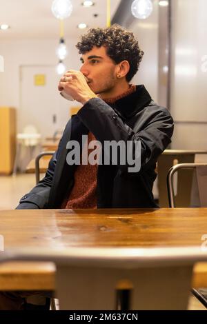 Giovane maschio in abiti caldi seduti in una moderna caffetteria mentre si ha una tazza di carta di bevande calde Foto Stock
