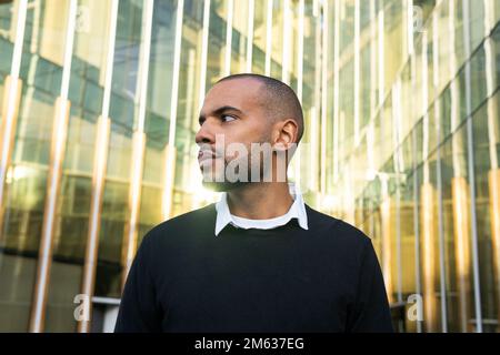 Basso angolo di serio imprenditore afro americano barbuto maschio in abbigliamento casual elegante in piedi contro contemporaneo edificio di vetro guardando lontano Foto Stock