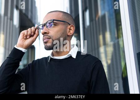 Basso angolo di etnia bearded maschio in occhiali e eleganti abiti casual in piedi vicino alla finestra del contemporaneo centro affari che guarda lontano Foto Stock