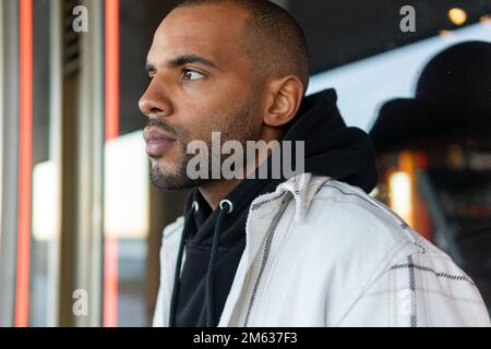 Vista laterale di un maschio afro-americano barbuto in felpa nera con cappuccio che si erge all'esterno dell'edificio moderno e che guarda lontano Foto Stock
