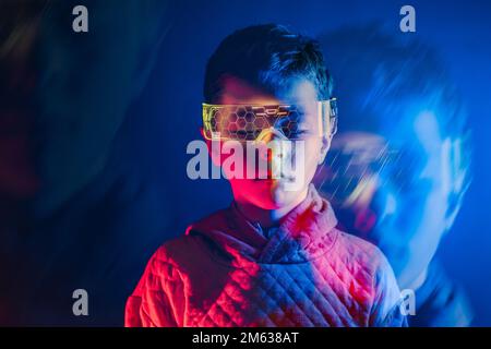 Bambino con occhiali intelligenti con occhi aperti che esplorano lo spazio virtuale mentre si trova in piedi nella stanza con illuminazione al neon blu Foto Stock